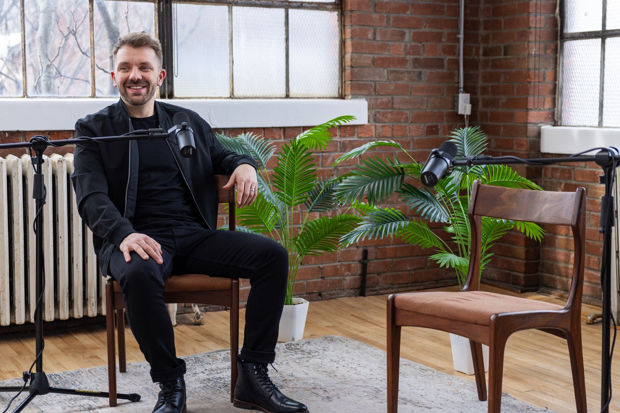 A guest seated in a brick-walled studio with a microphone on a stand, plants and a camera nearby