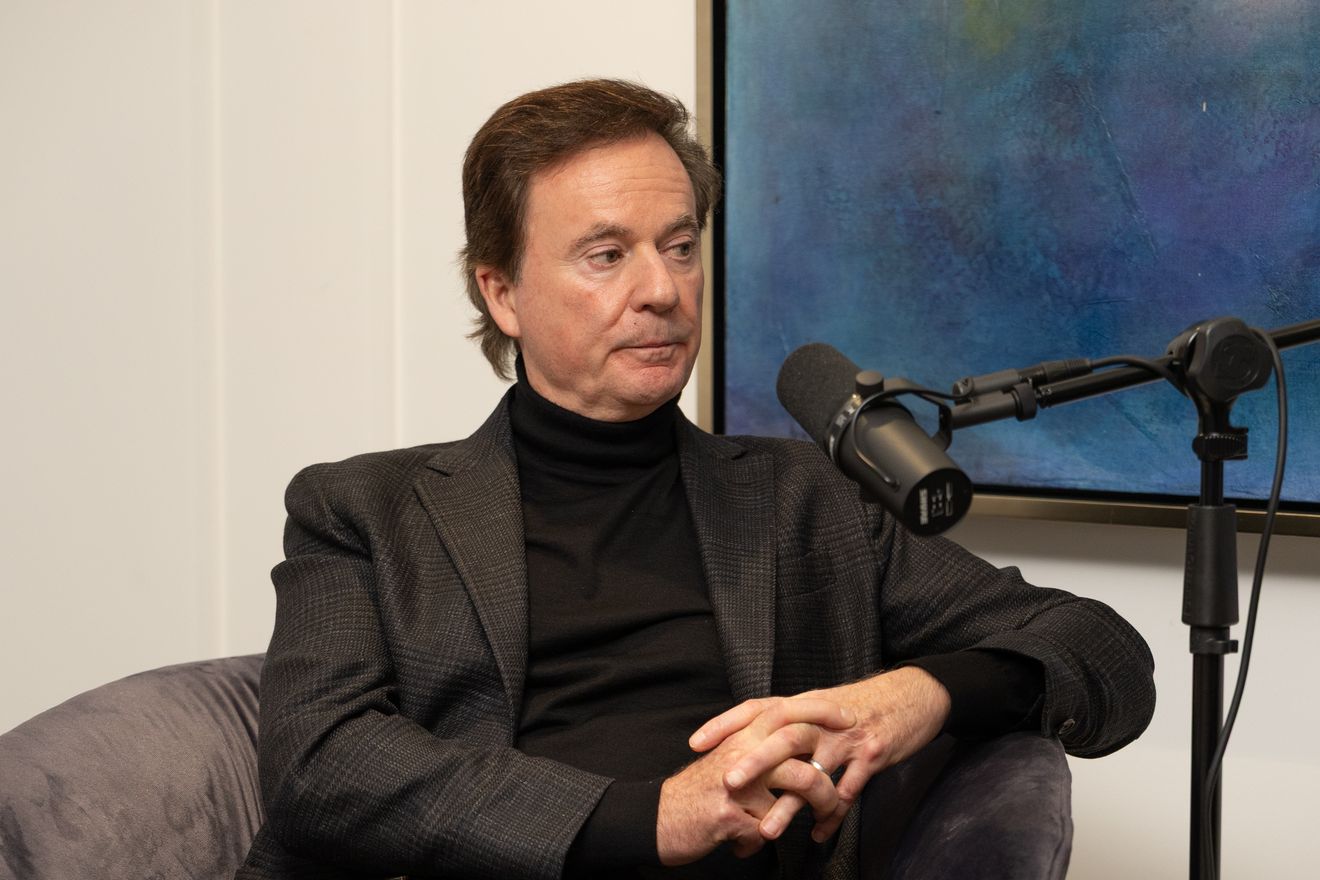 A guest in a dark jacket seated in a grey armchair beside a microphone on a boom arm