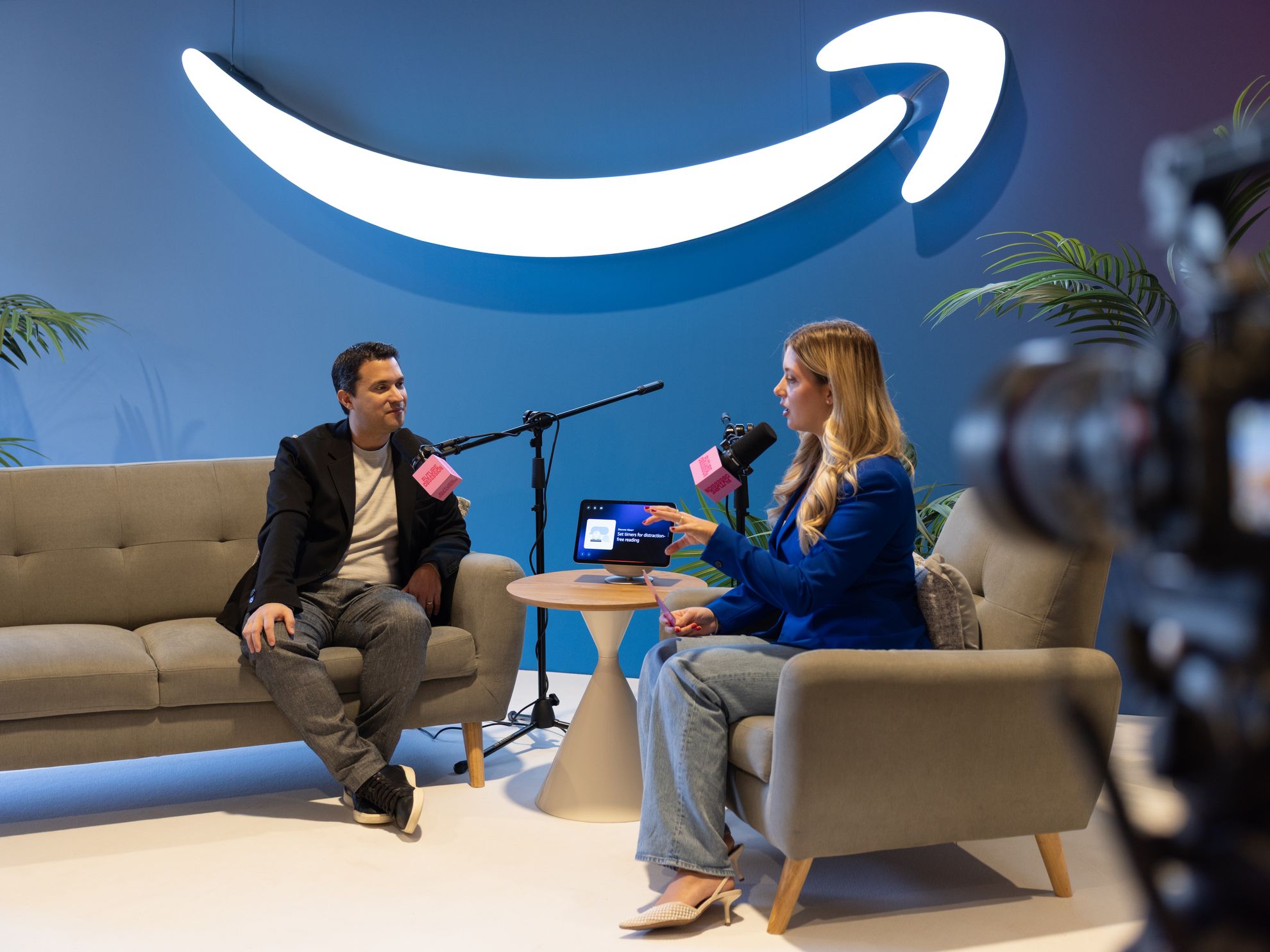 A host and guest talking on a sofa set, a laptop on the table between them and a camera in the foreground