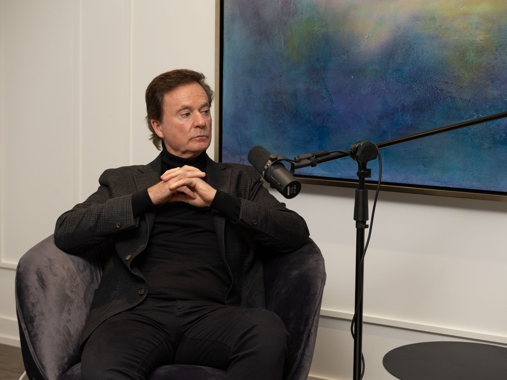 A guest seated with hands folded in front of a large painting, a microphone on a boom arm beside them