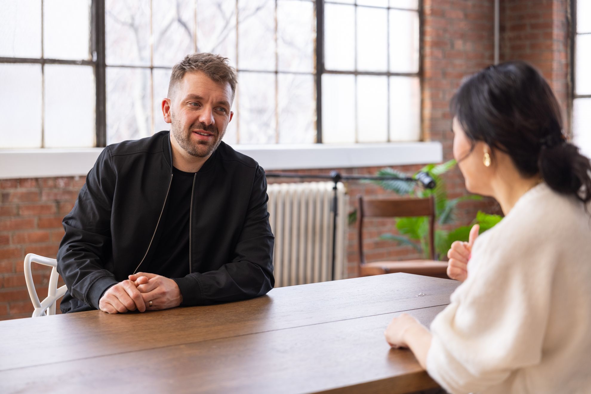 A guest laughing at a table in a brick-walled studio, facing the host across from them