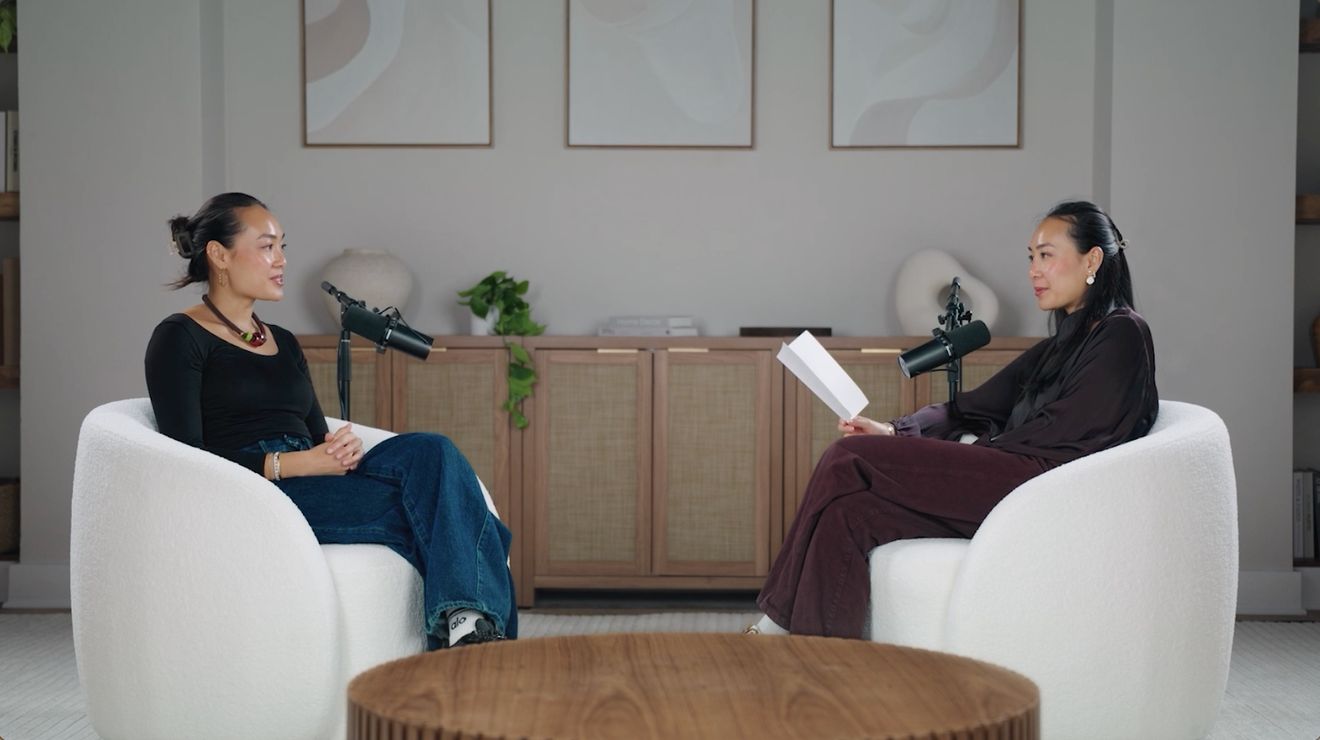 Wide view of two women facing each other in white armchairs, each at a microphone