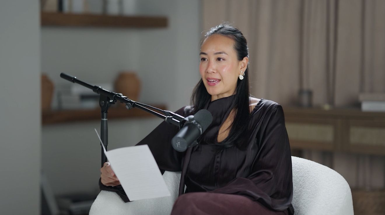 A woman in a dark satin top reading from a sheet of paper at a podcast microphone