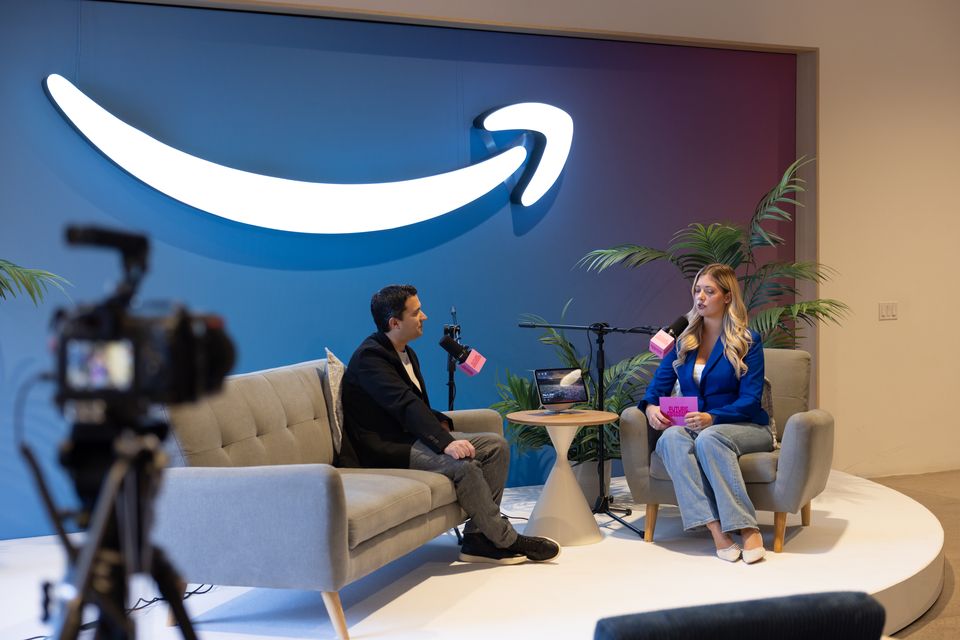 Host and guest on a sofa in front of a large logo wall, with a camera on a tripod in the foreground
