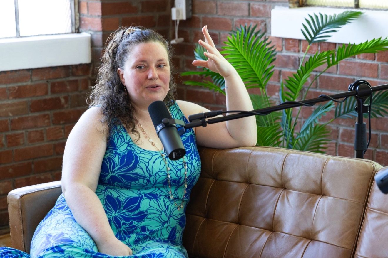 Guest in a blue floral dress speaking at a microphone on a leather couch in the loft