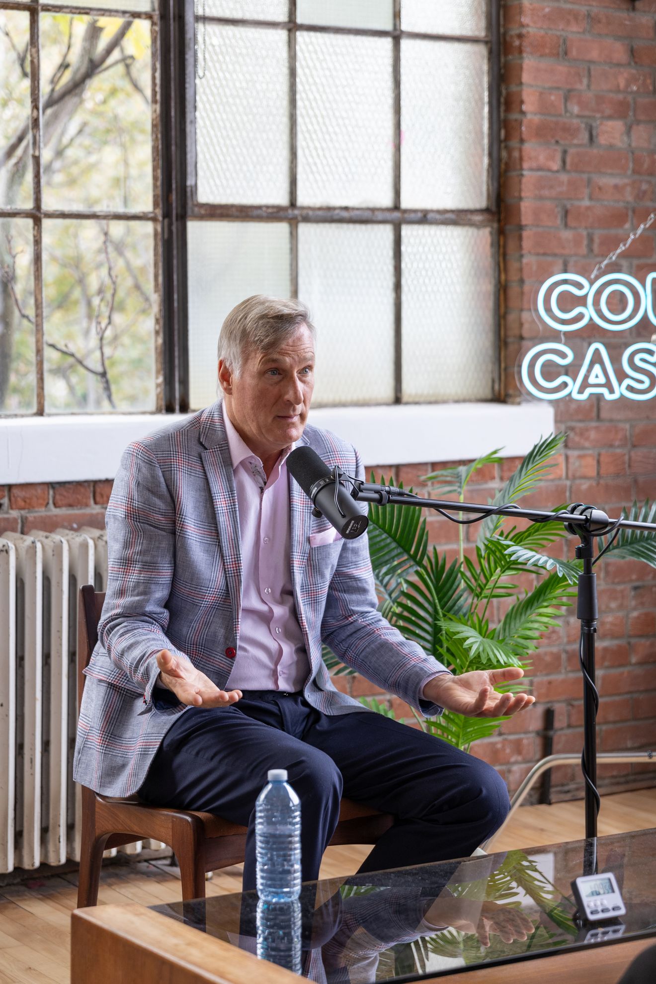 Guest in a grey jacket seated at a microphone beside a big window in the brick loft