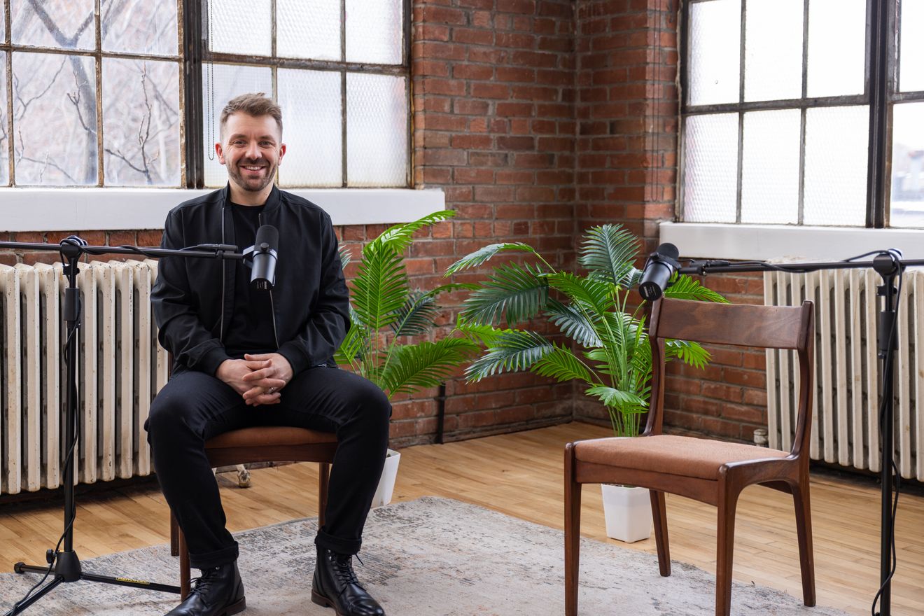 Guest seated in a wooden chair at a microphone in the brick loft, with plants and windows behind