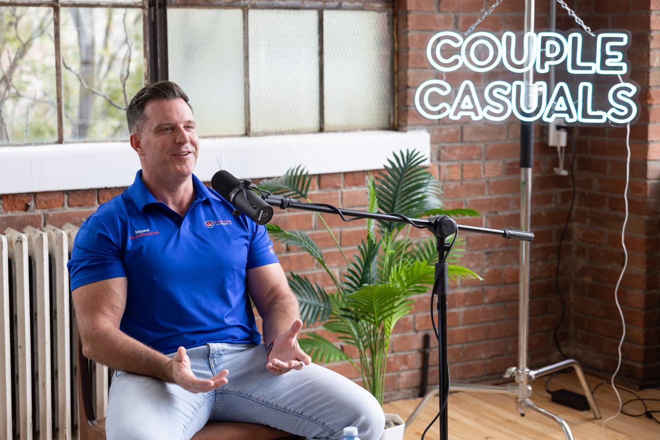 Guest in a blue shirt speaking at a microphone beside a radiator and plants in the brick loft