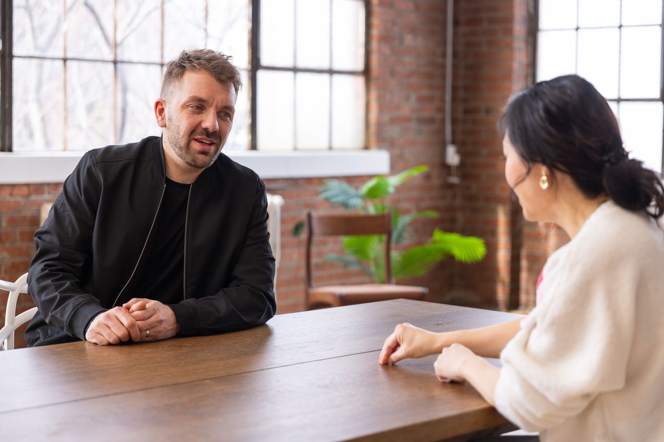 Host and guest at a wooden table in front of brick and windows