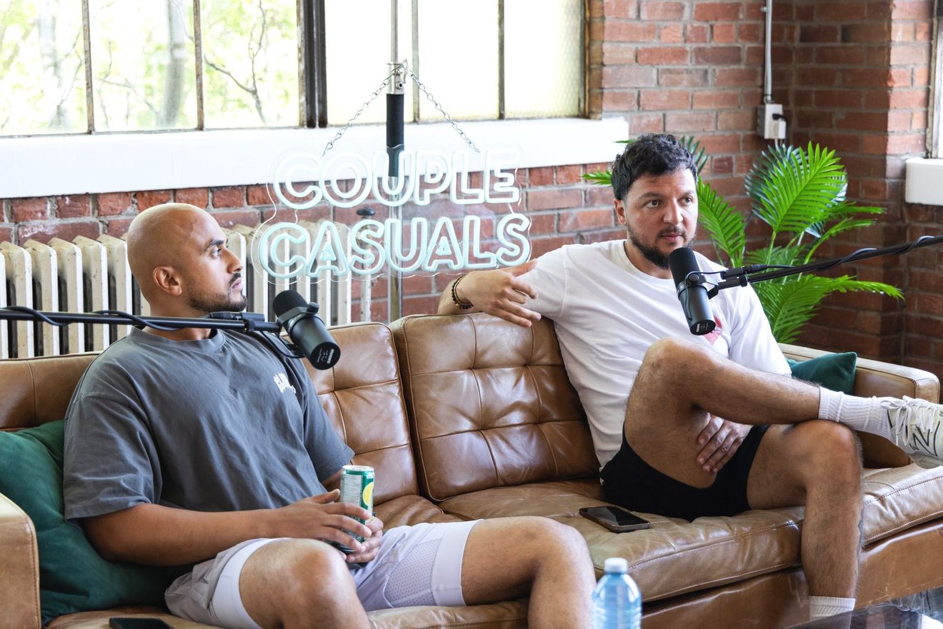 Two guests talking on a tan leather couch, each with a microphone, in front of brick and a radiator