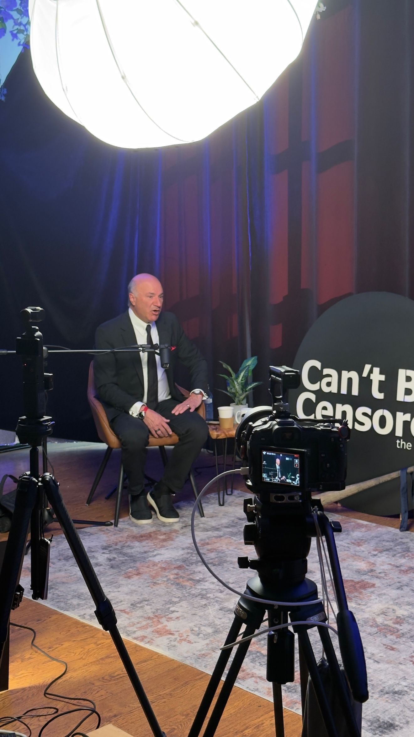 Wide view of a dark set with blue curtains, a round sign and cameras, with a guest seated at the back