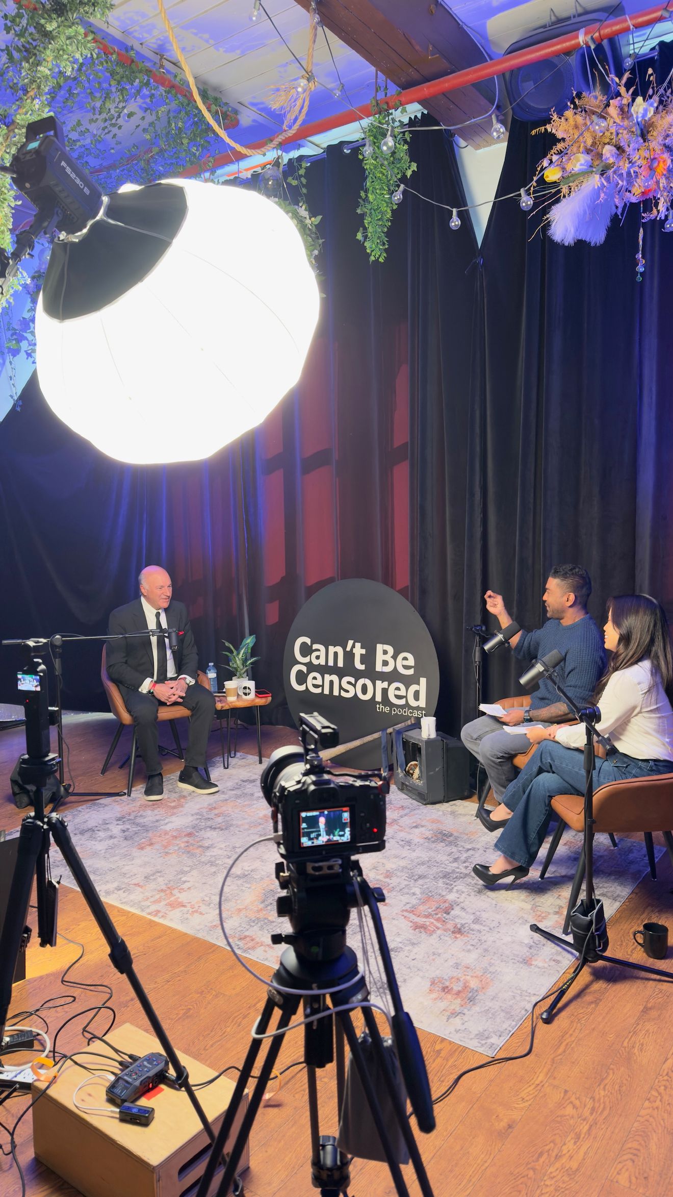 Dark set with blue curtains, a softbox light and cameras, with a host and guests seated in the distance