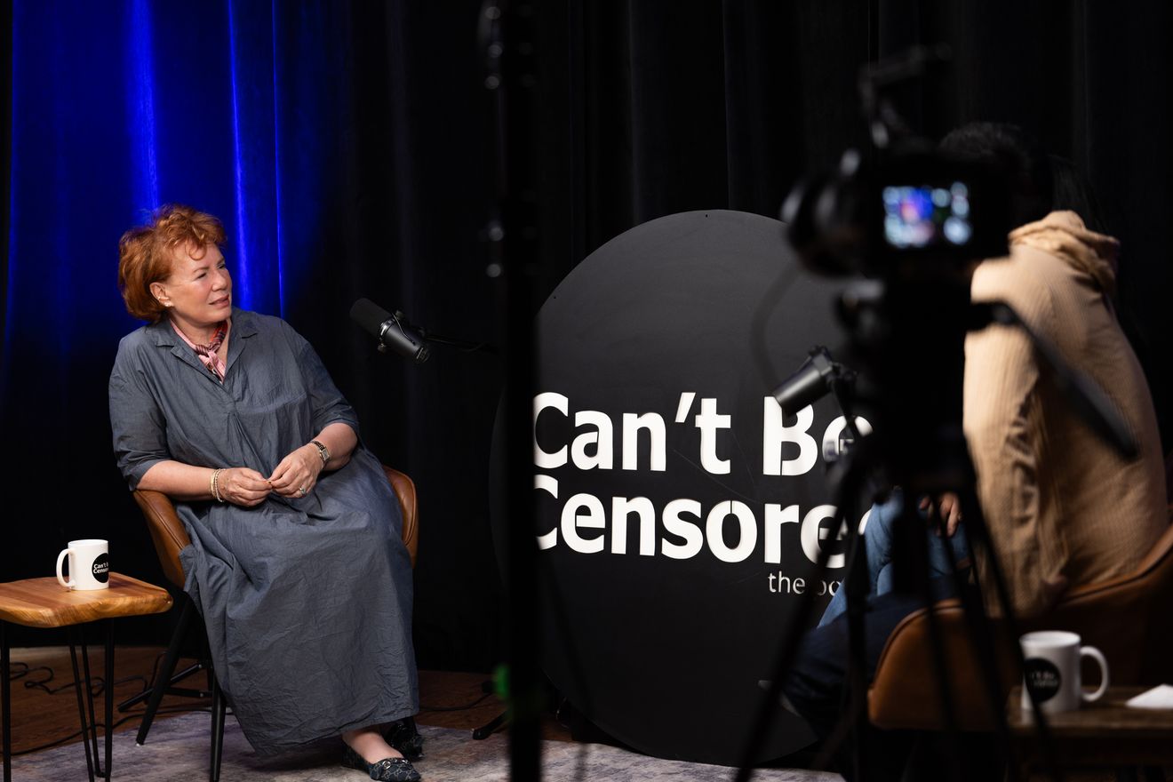 Guest seated in a leather chair on a dark set with blue curtains, with a large round sign beside them