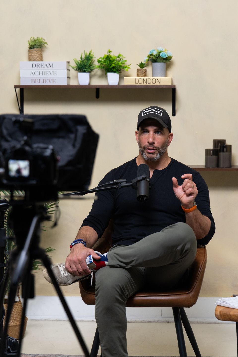 Guest speaking in front of a tan wall with shelves, beside a camera