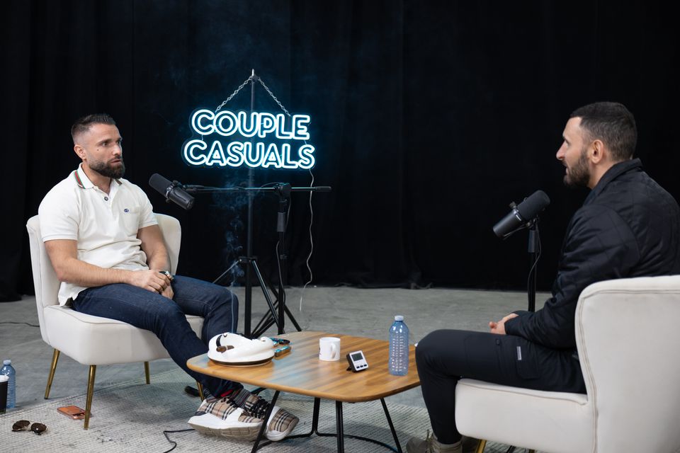 Two hosts on a dark set with a neon sign and a coffee table