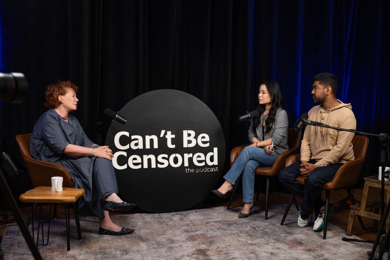 Three people in conversation at microphones on a podcast set with a round logo sign against dark curtains