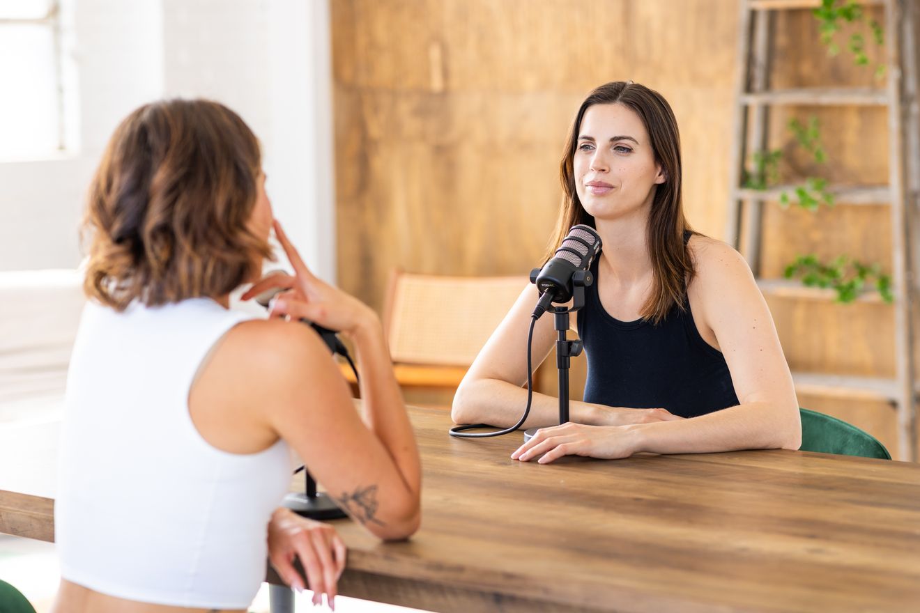 Two people in conversation at a wooden table, one speaking into a podcast microphone