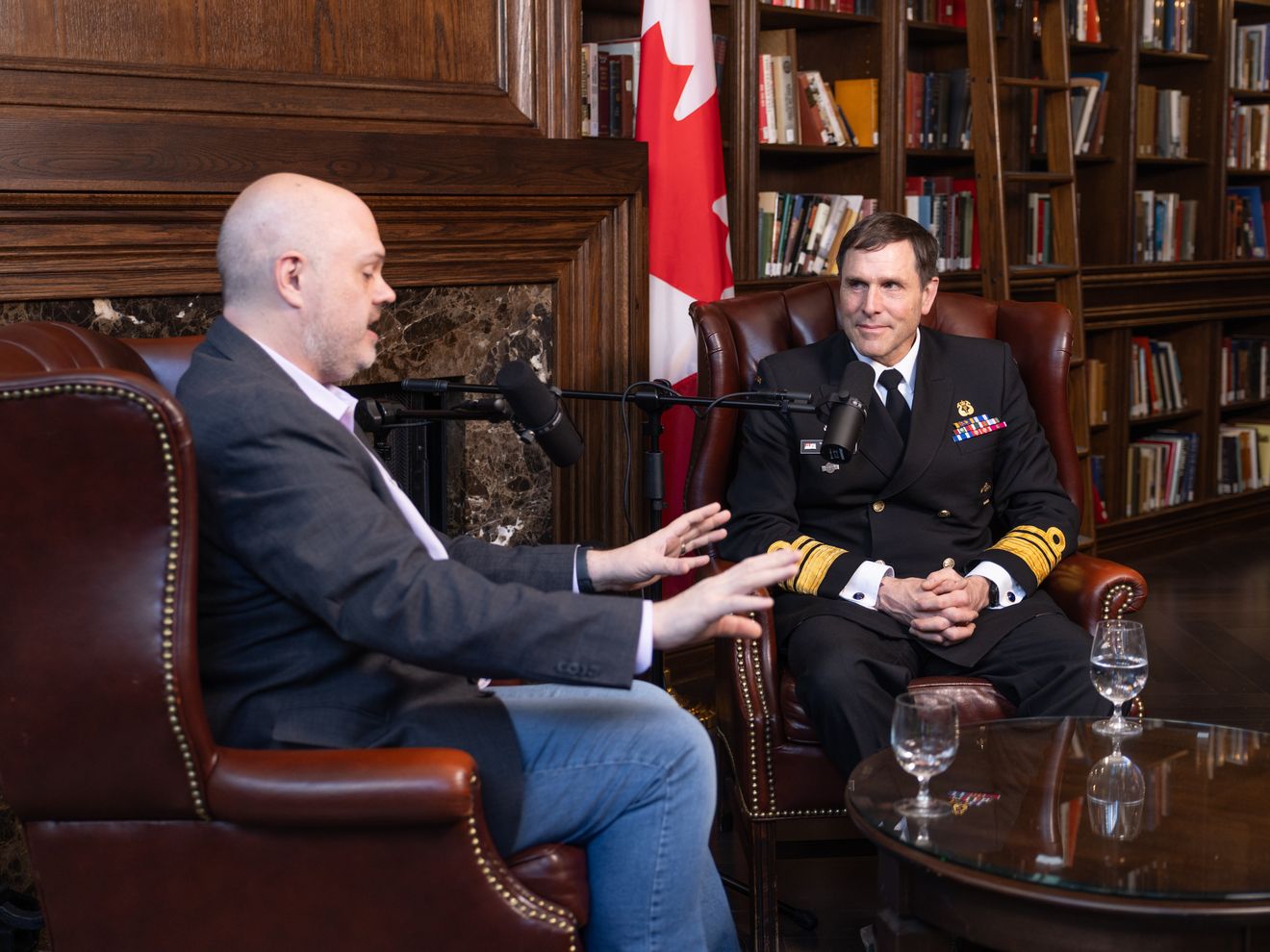 Two men in leather armchairs in a wood-panelled library with bookshelves and a Canadian flag, being filmed for a podcast