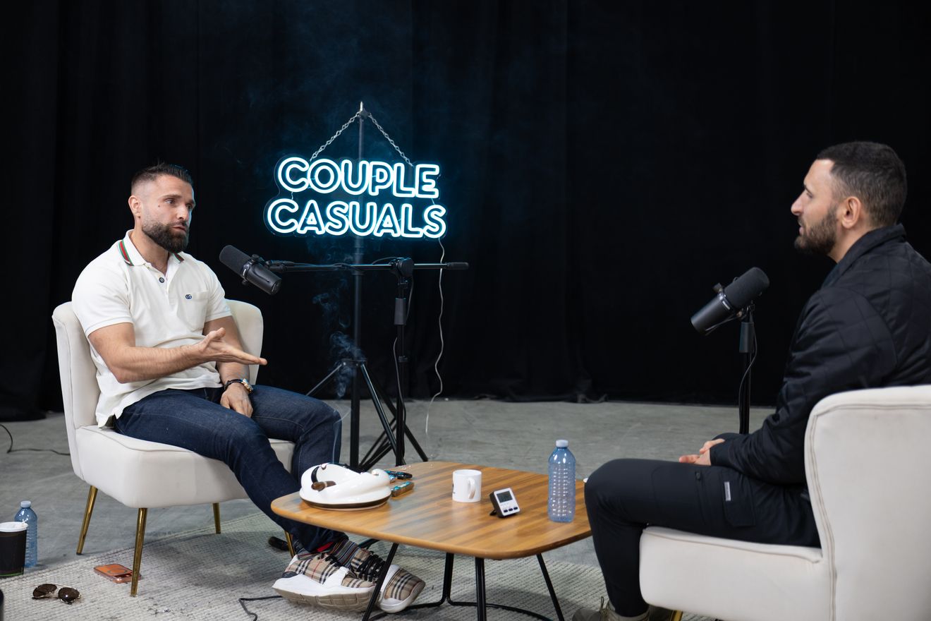 Two men recording a podcast on a dark set with a glowing neon sign and a microphone in front of each