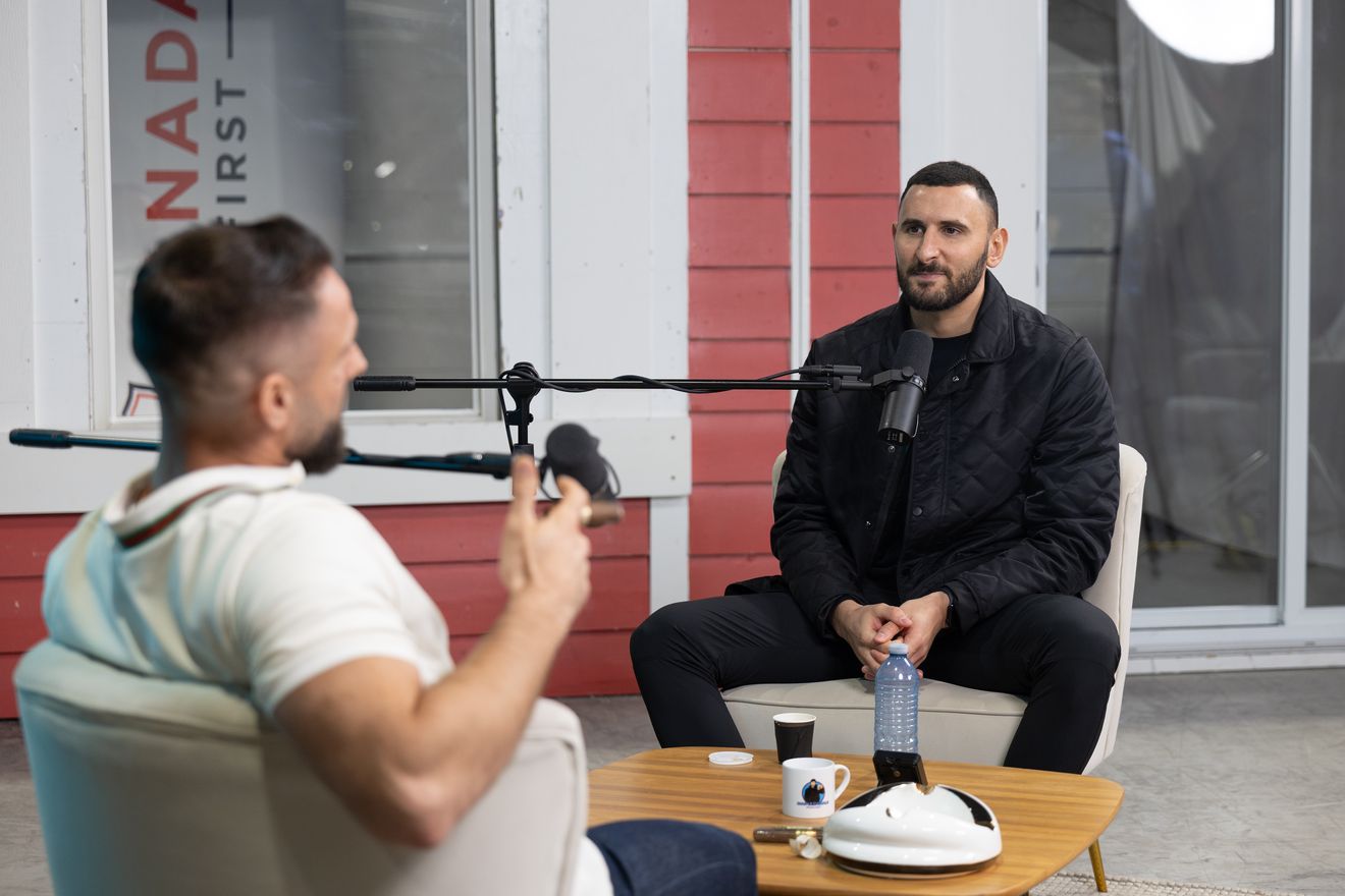 Two men in conversation at microphones on a small table, with a red-painted house behind them, filmed on location